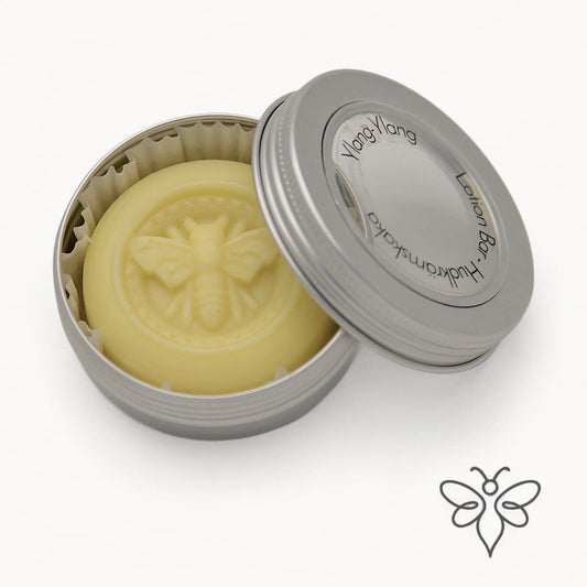 EneboBee Hudkrämskaka – Lotion Bar med bivax i doften Ylang-Ylang i en öppnad metallburk med locket lutad mot en silverfärgad burk.